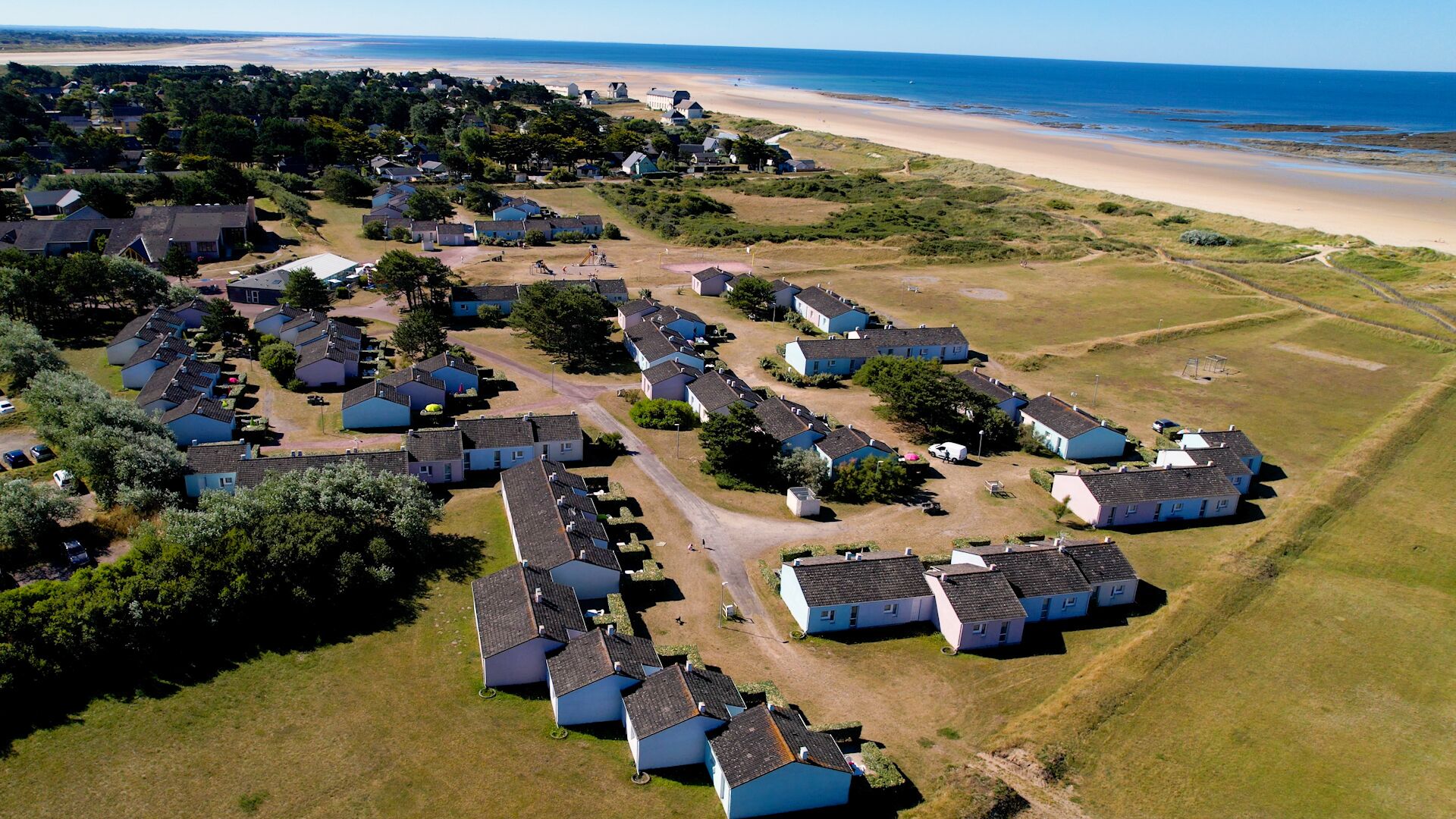 Vista aérea de una zona residencial costera con casas alineadas en filas, una playa al fondo y cielo despejado.