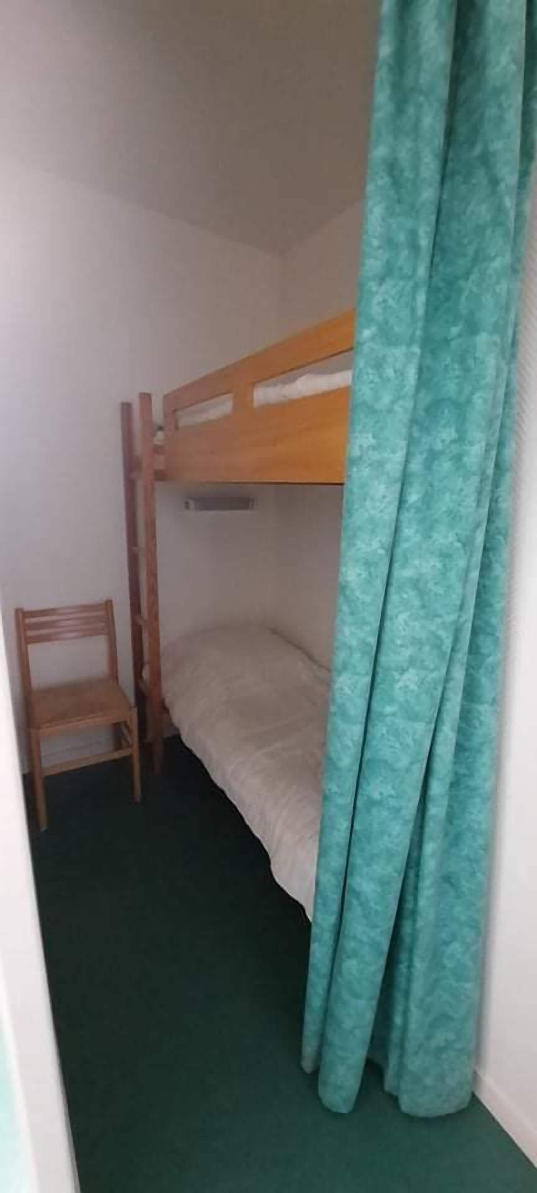 Una habitación pequeña y funcional con una cama litera, una silla de madera y una cortina color turquesa.
