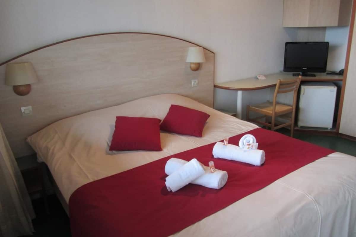 Une chambre d’hôtel avec un lit, des oreillers rouges, une couverture rouge et deux serviettes enroulées. On y trouve un bureau avec un ordinateur et une chaise.
