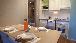 Un coin repas avec une table en bois mise pour quatre personnes, comprenant des assiettes, des verres et des couverts. Une bouteille contenant un liquide orange et un vase bleu sont posés sur la table. La pièce est équipée de meubles blancs, d’un four à micro-ondes et d’un lave-vaisselle.