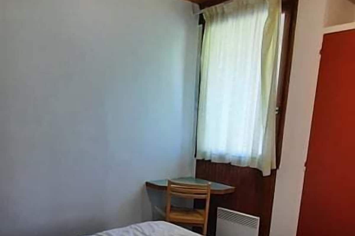 Une chambre avec un lit simple, un plafond en bois, une petite table, une chaise et une fenêtre avec des rideaux.