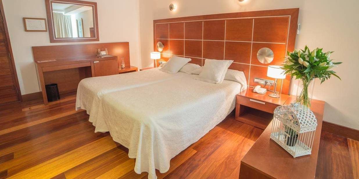 Habitació d'hotel amb un llit gran, una taula de nit de fusta, un mirall, una cambrera amb una làmpada i una planta decorativa.