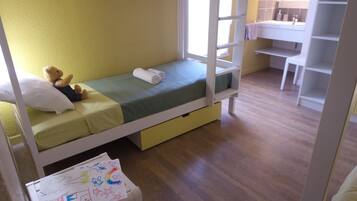 A single bed with a green mattress, a teddy bear, and a white pillow. There is a small desk with a chair and a mirror above it. The room has wooden flooring and yellow walls.