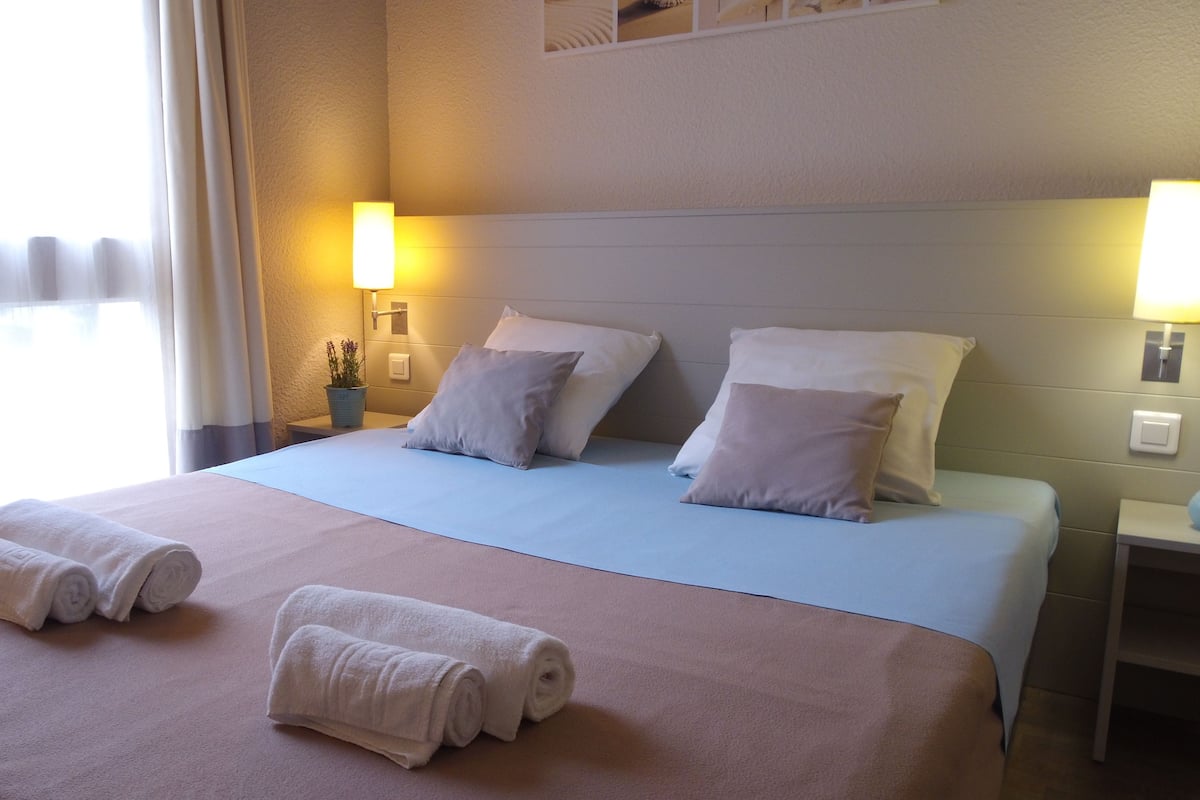 Quarto de hotel com uma cama grande, duas luminárias laterais e uma janela com cortinas.