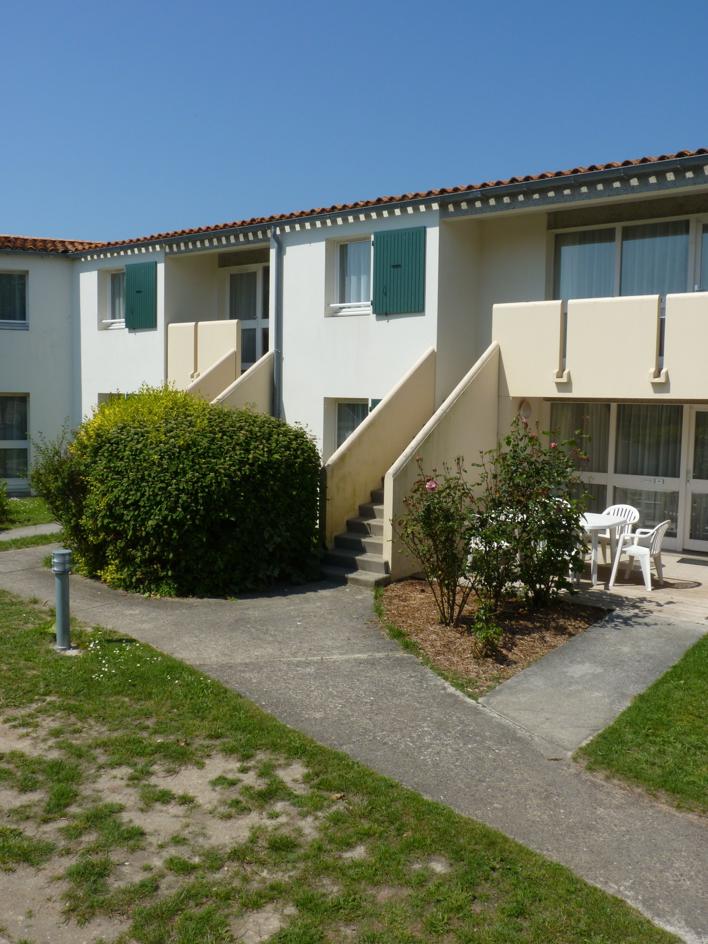 A building with a staircase leading to the entrance, a small garden area with a bush and a flower, and a seating area with chairs.