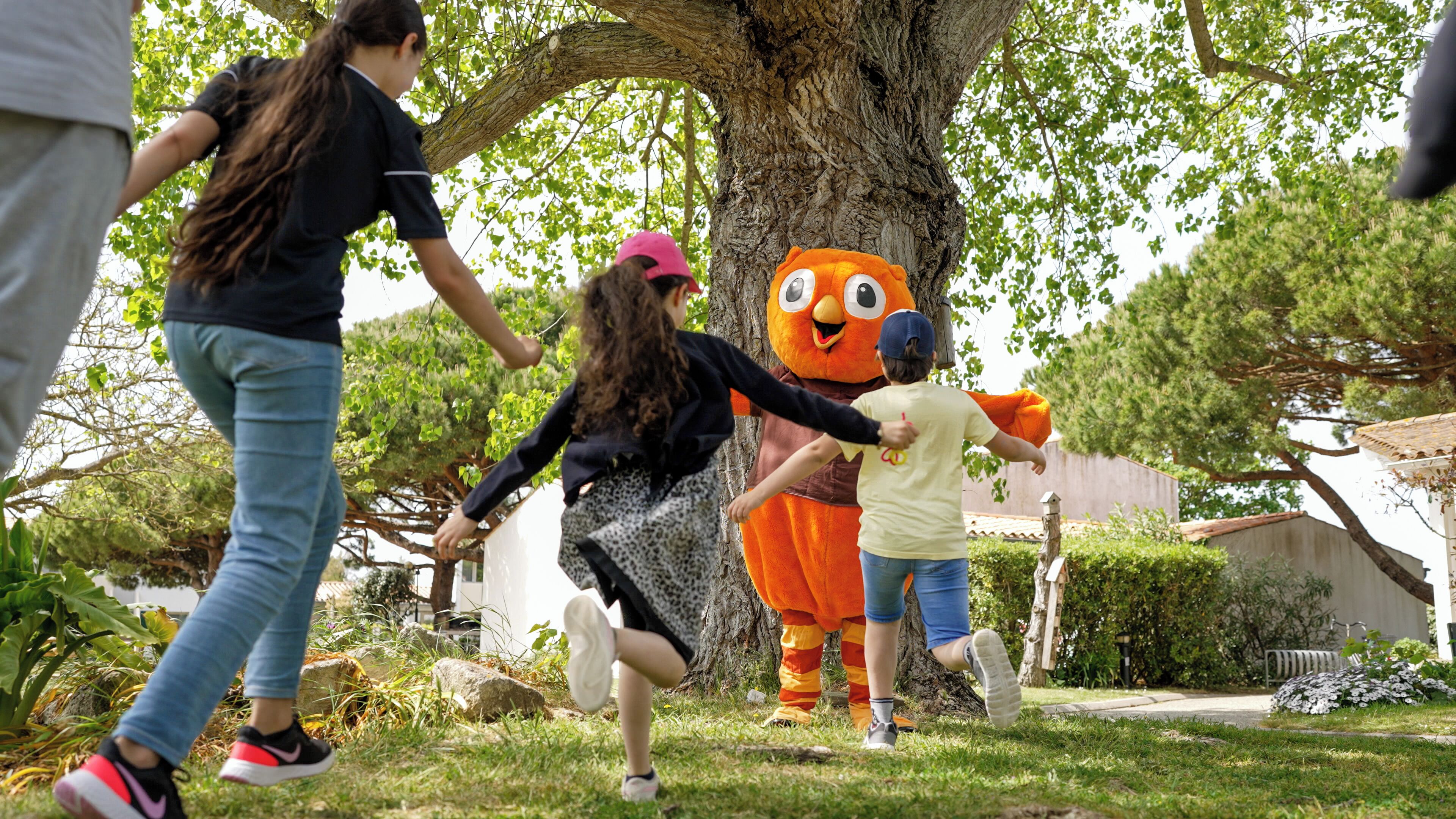 Un groupe de personnes interagissant avec une grande mascotte orange dans un espace extérieur résidentiel.