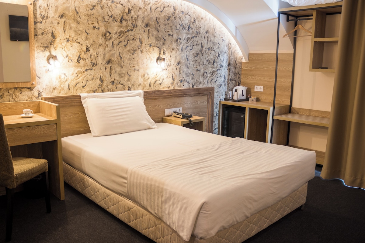 A hotel room with a marble wall, a bed with a quilted mattress, a wooden nightstand, a chair, and a small table with a cup.
