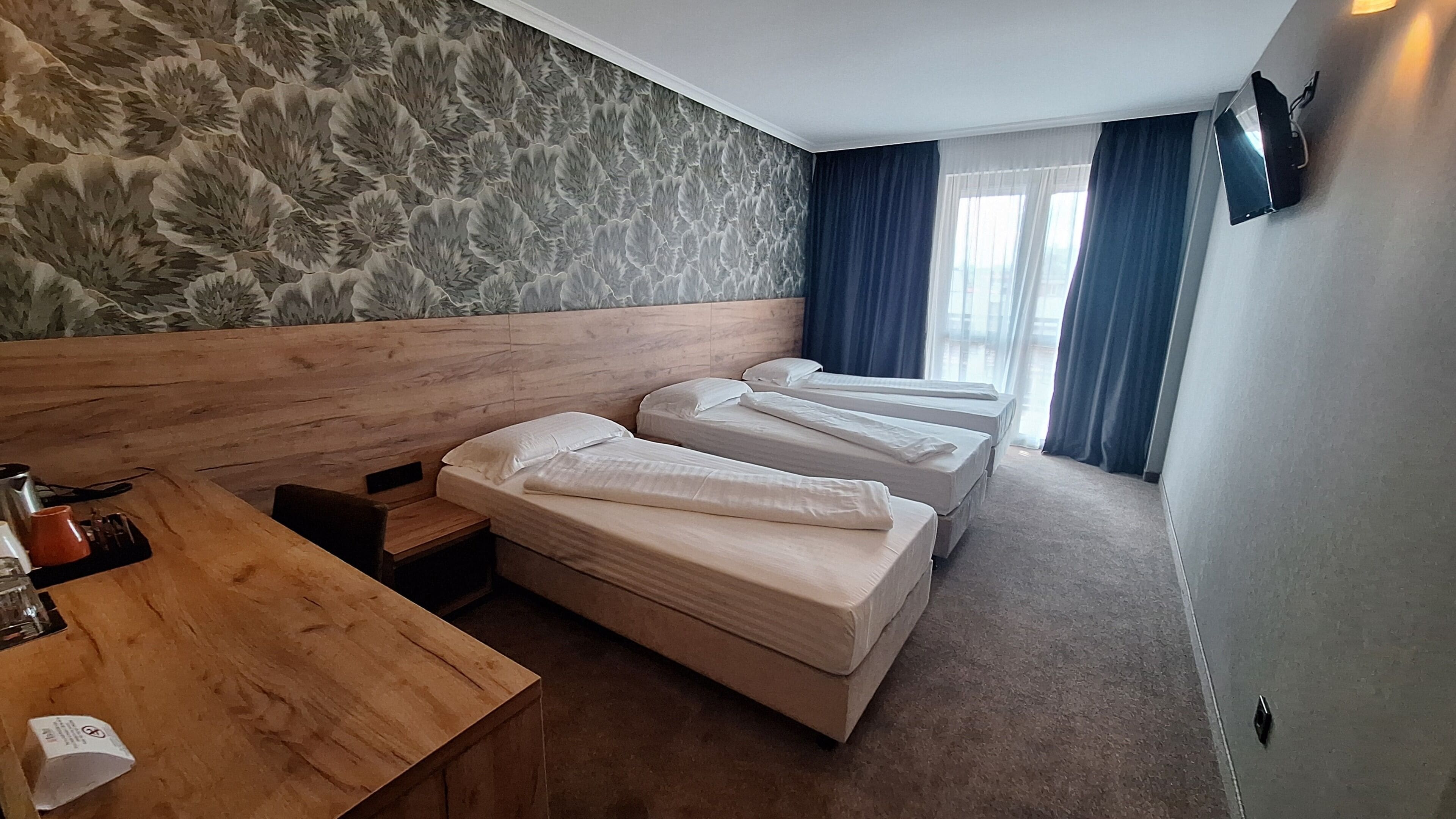 A hotel room with a wooden headboard, three beds, a wooden desk, and a patterned wallpaper.