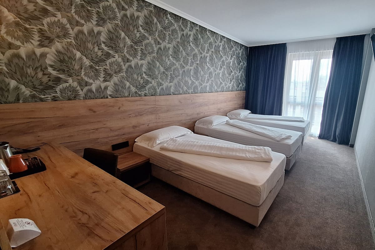 A hotel room with a wooden headboard, three beds, a wooden desk, and a patterned wallpaper.