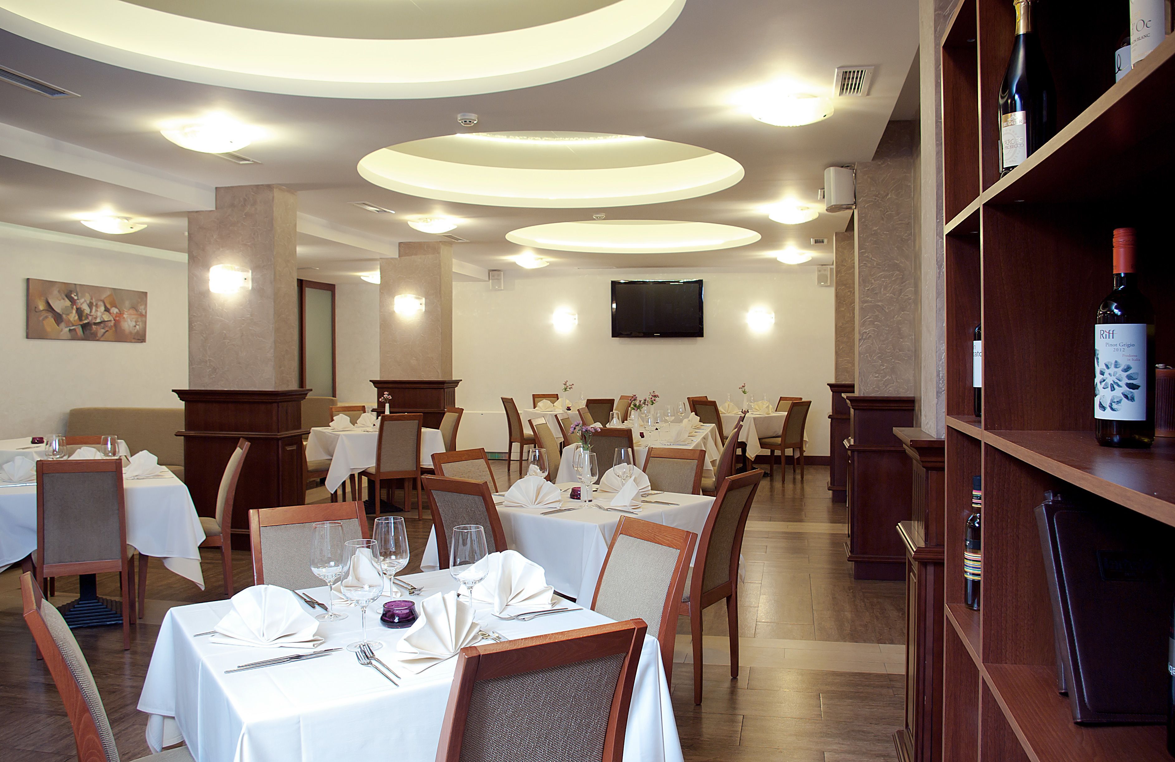 A restaurant with round tables set for dining, a wine bottle on a shelf, and a television on the wall.