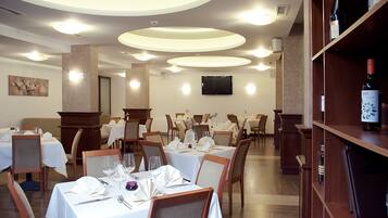 A restaurant with round tables set for dining, a wine bottle on a shelf, and a television on the wall.