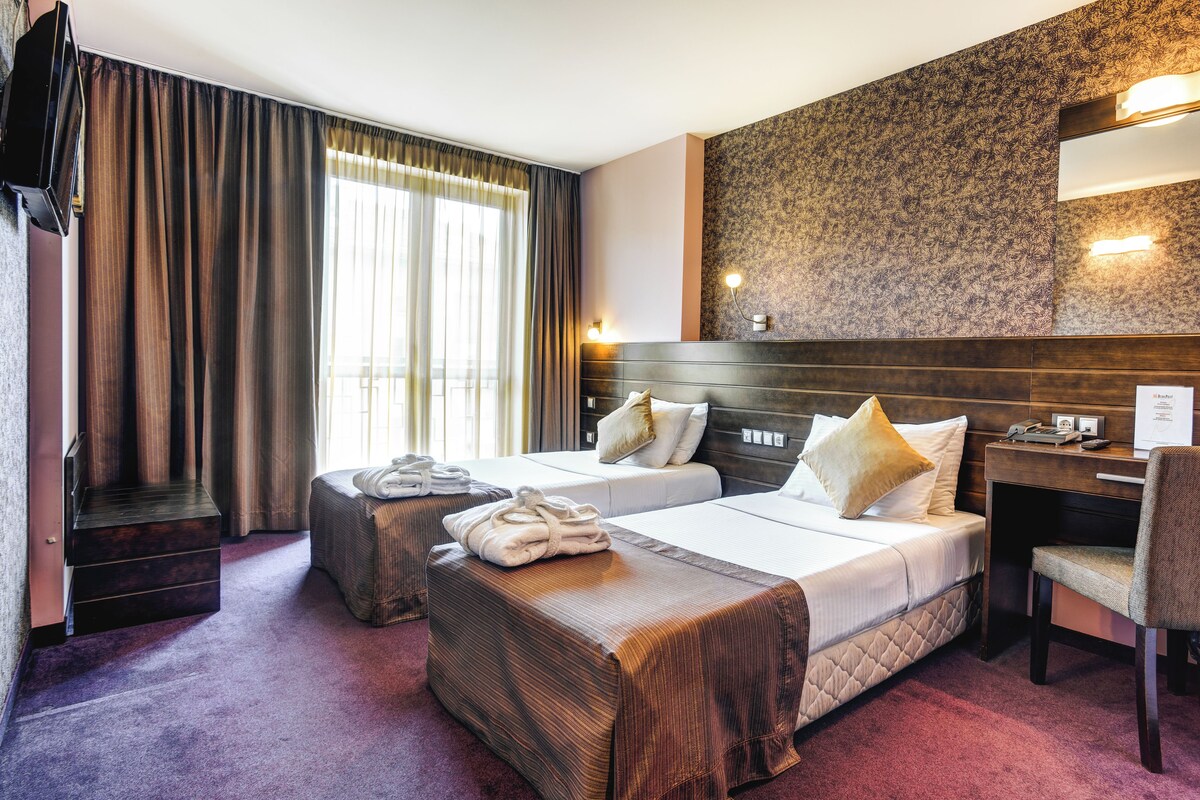 A hotel room with two beds, a desk, and a chair. There is a TV mounted on the wall, and a window with curtains.