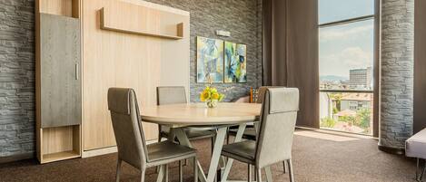 A modern room with a round table, four chairs, a wooden cabinet, and a large window offering a city view.