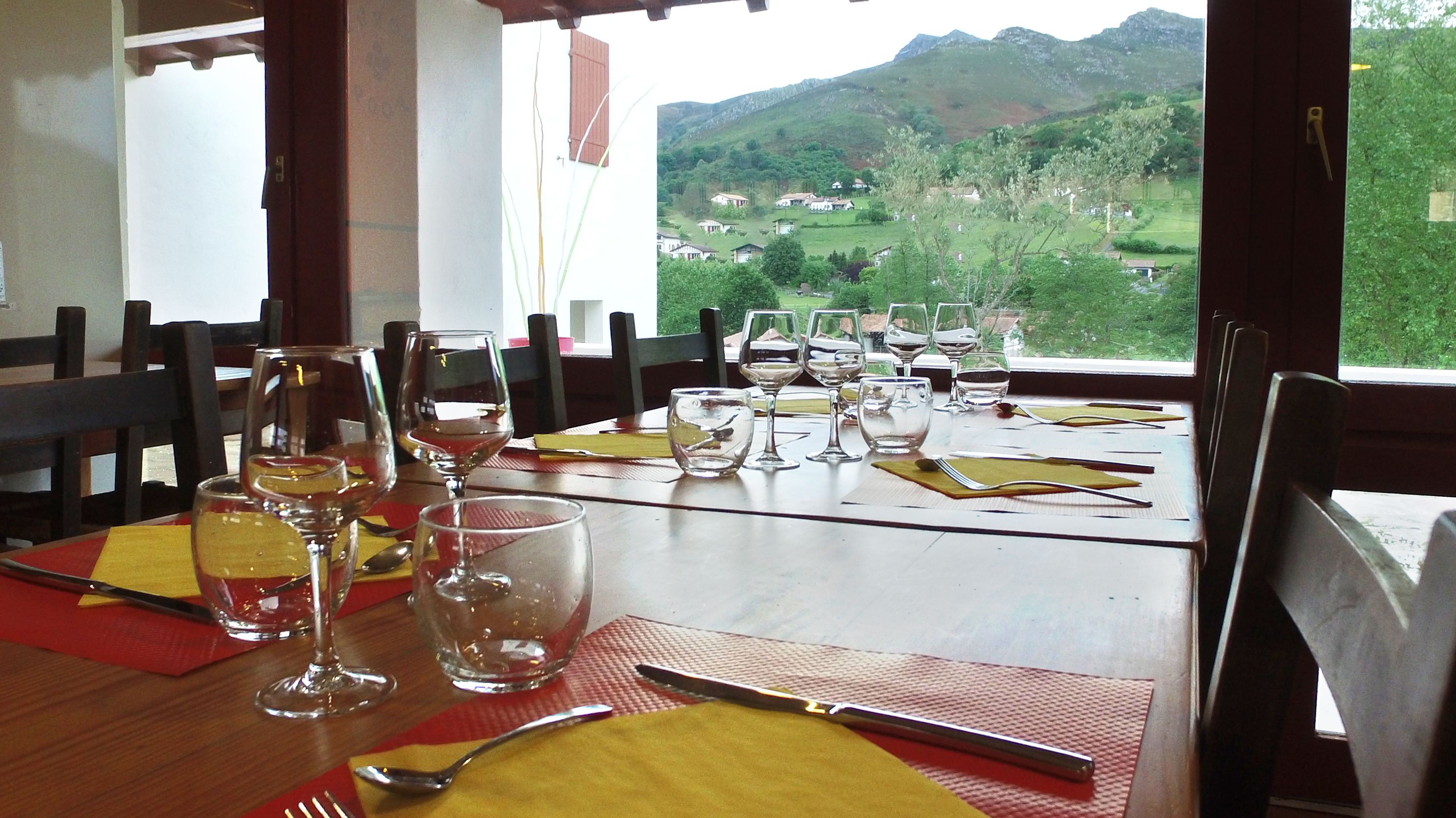 Une table à manger dressée avec des verres à vin, des couverts et des serviettes, offrant une vue pittoresque sur des maisons et des collines.