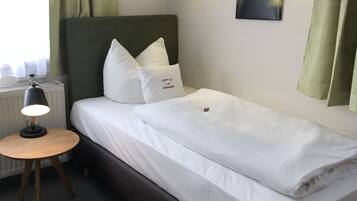 Ein ordentlich bezogenes Bett mit weißen Leinen und Kissen, ein runder Holfnachtisch mit Lampe, eine Heizung und ein Gemälde von einem Wald an der Wand.