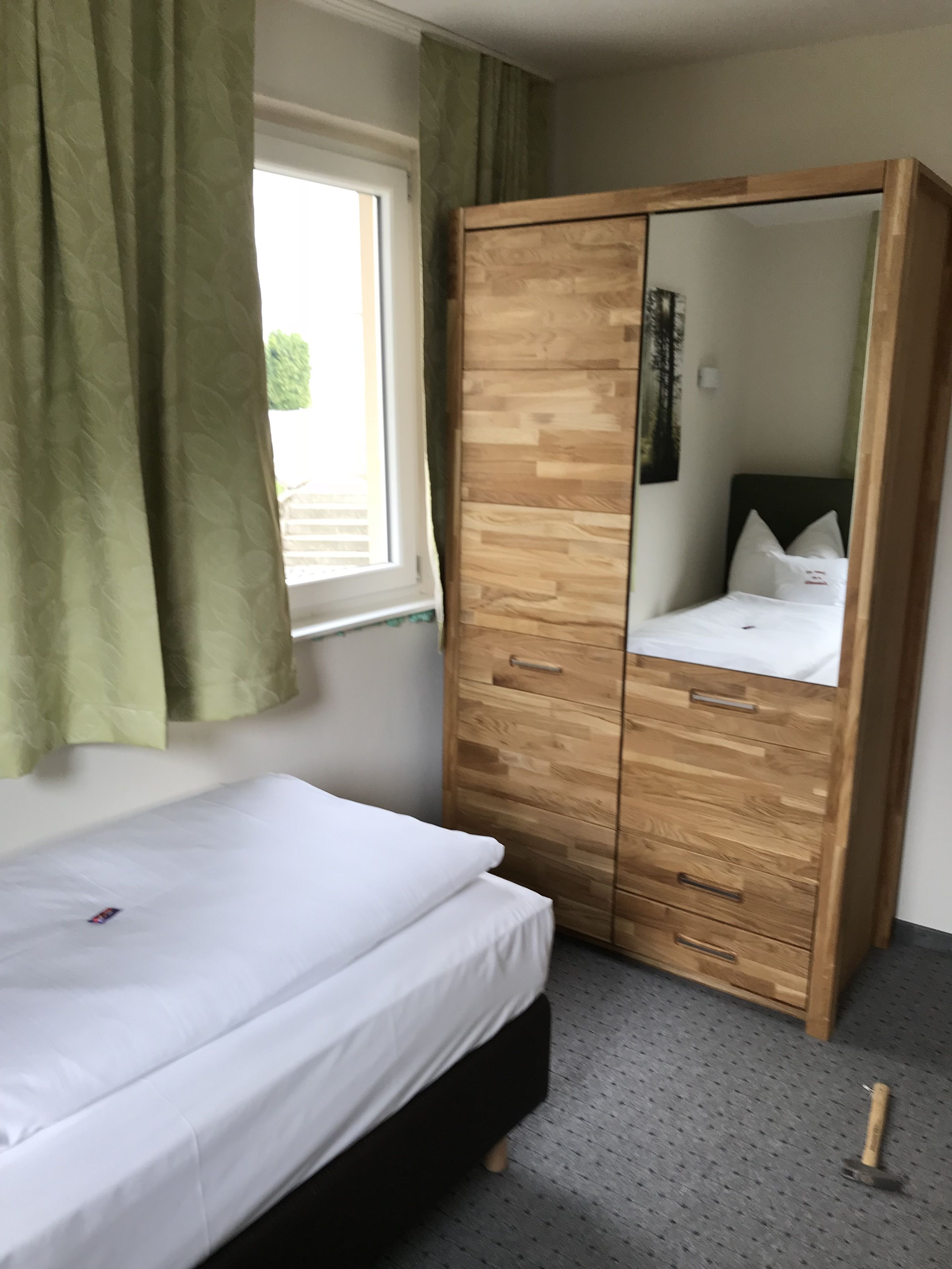 A bedroom with a bed, a wooden wardrobe, a window with curtains, and a mirror.