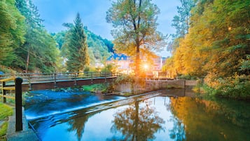 A calm river with a bridge, surrounded by lush greenery and a building with a blue roof in the background.