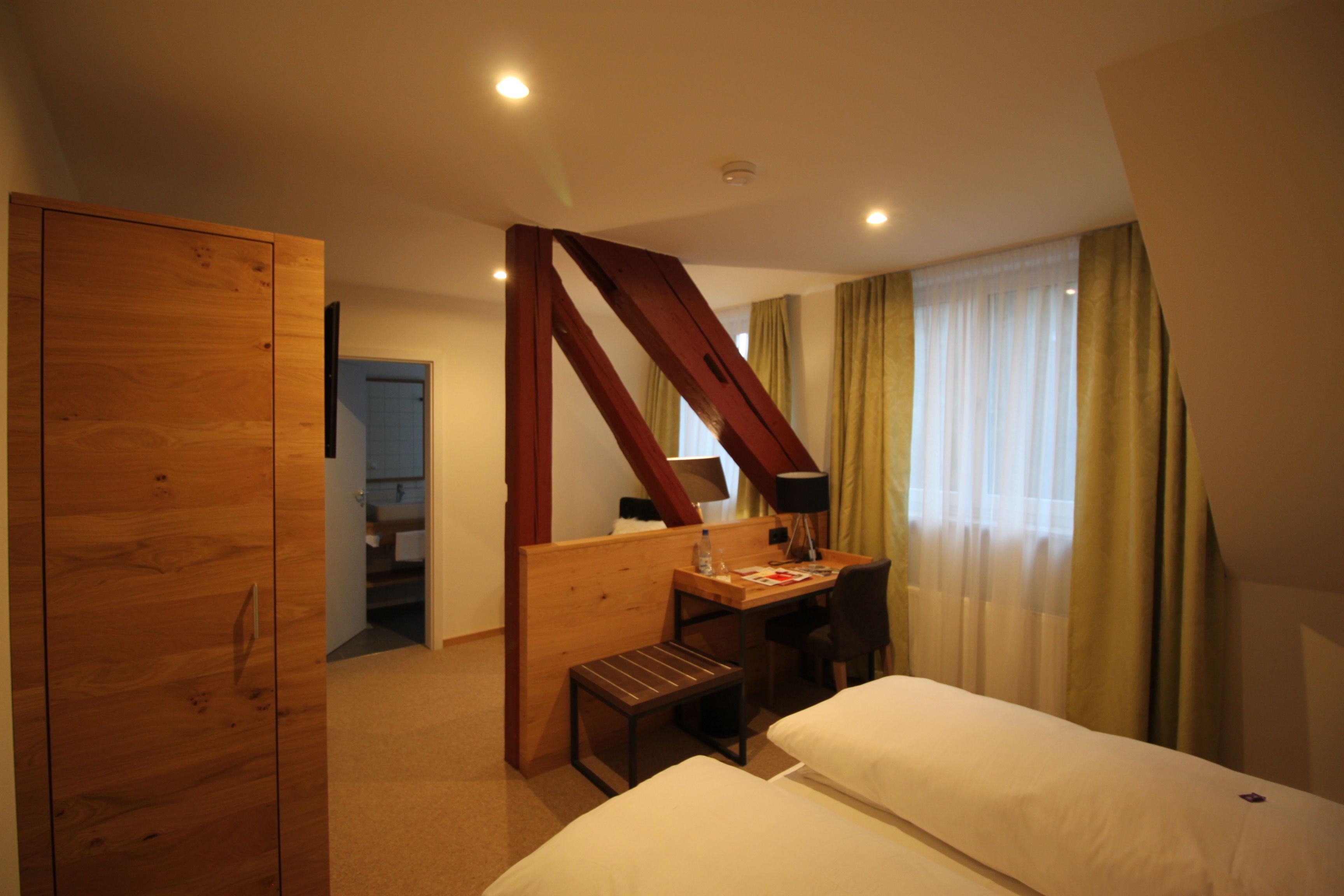 A hotel room with a wooden wardrobe, a desk with a chair, a small bench, two beds, and a window with curtains.