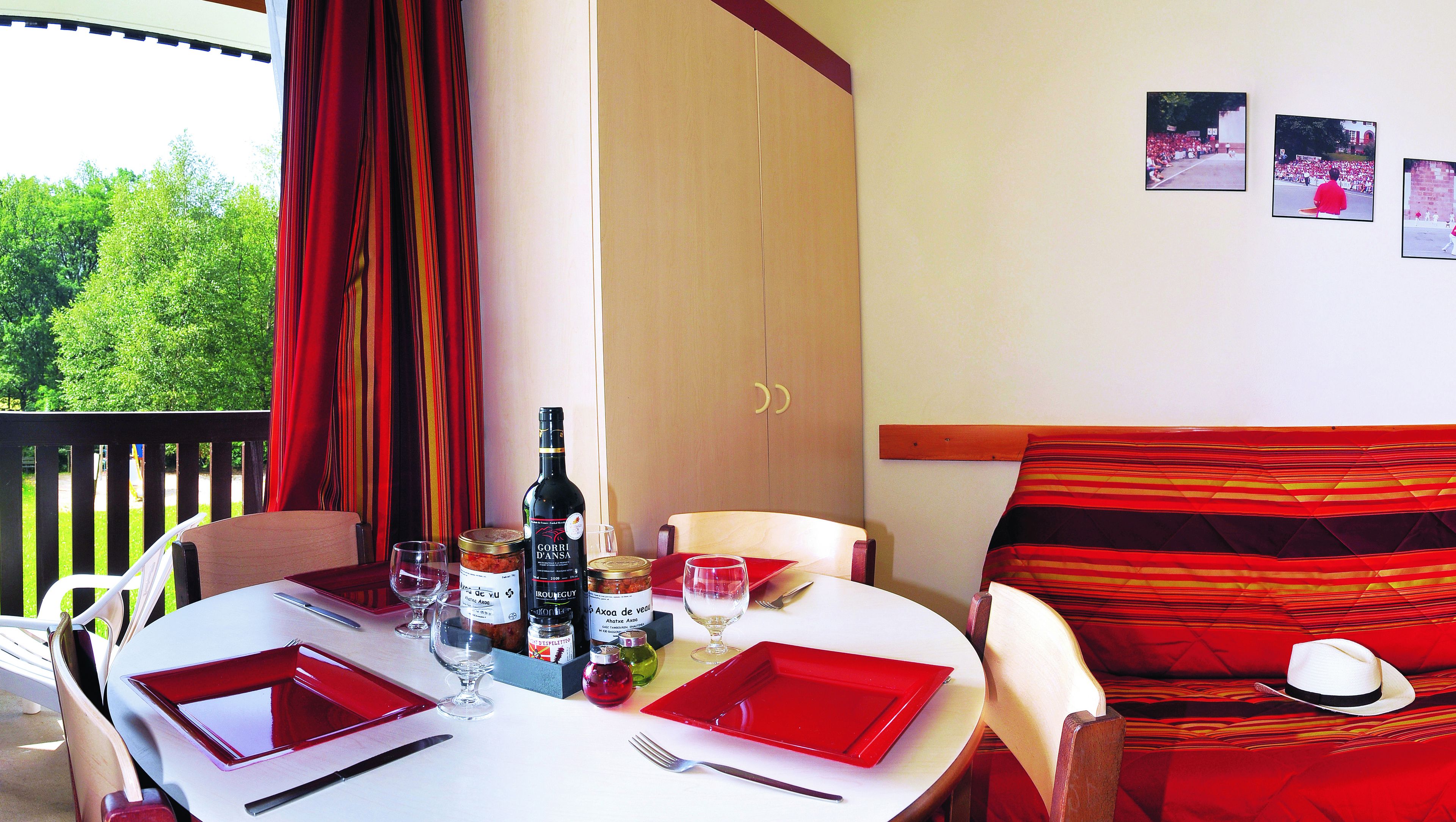 Mesa de comedor dispuesta con vajilla roja, botellas de vino y copas, con vistas a zonas verdes al exterior.