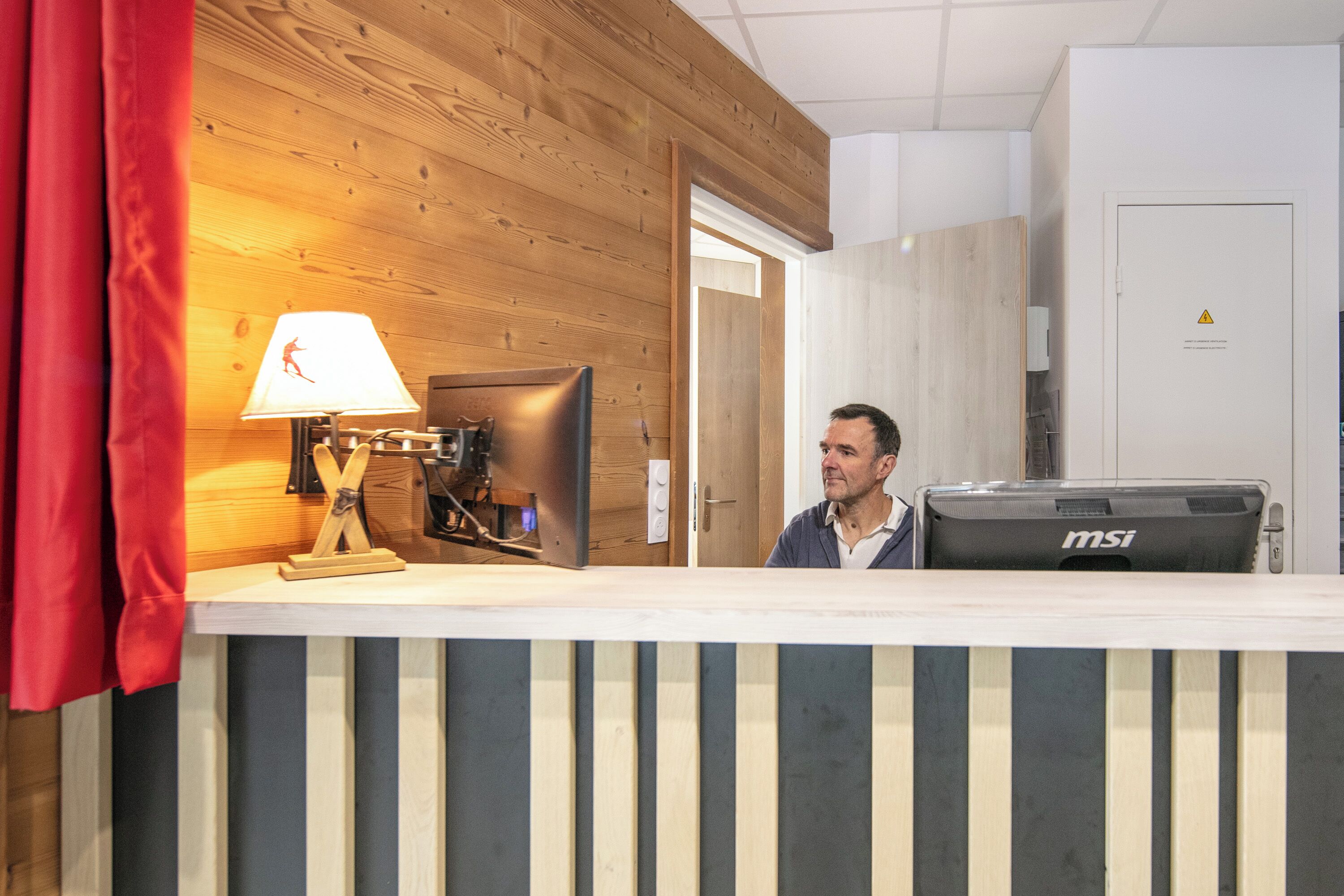 A reception desk with a lamp, a computer, and a suitcase labeled "msi" in a room with wooden paneling and a door in the background.