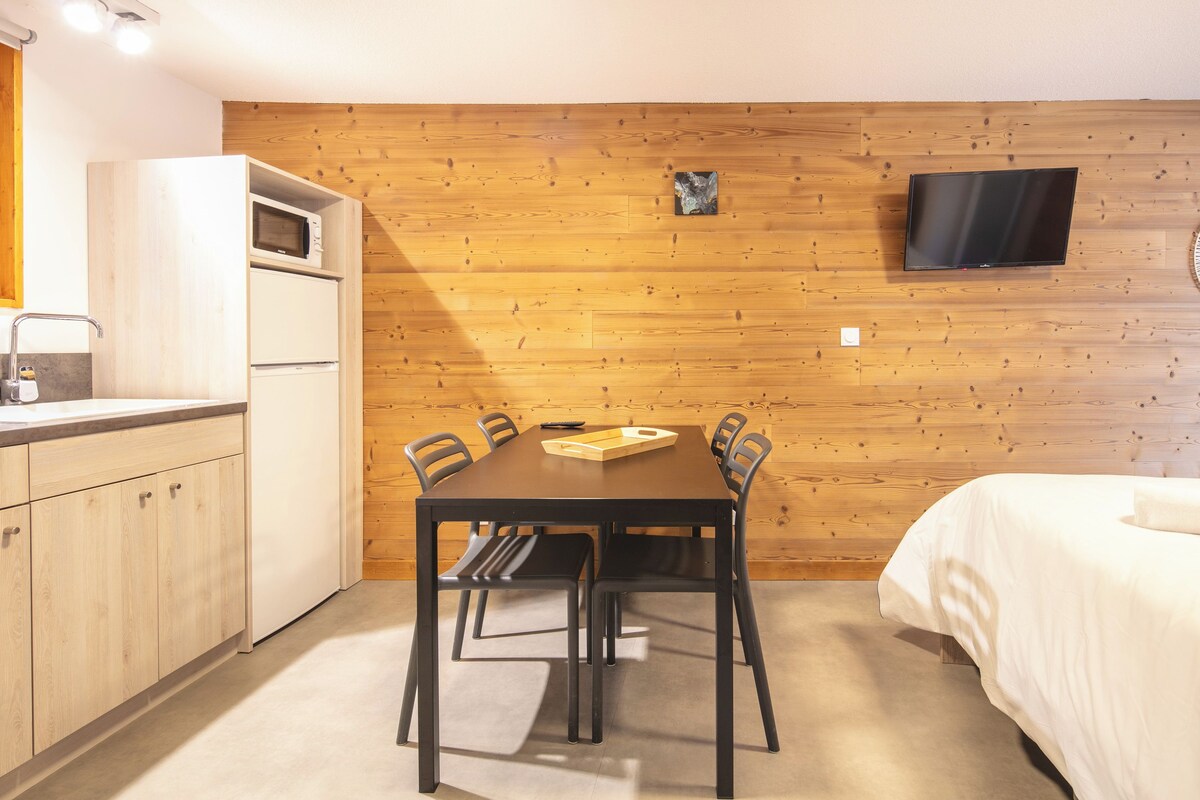 Une pièce compacte avec un mur en bois, un téléviseur à écran plat fixé au-dessus d’une table en bois avec des chaises, une petite kitchenette équipée d’un four à micro-ondes et un lit avec du linge de lit blanc.