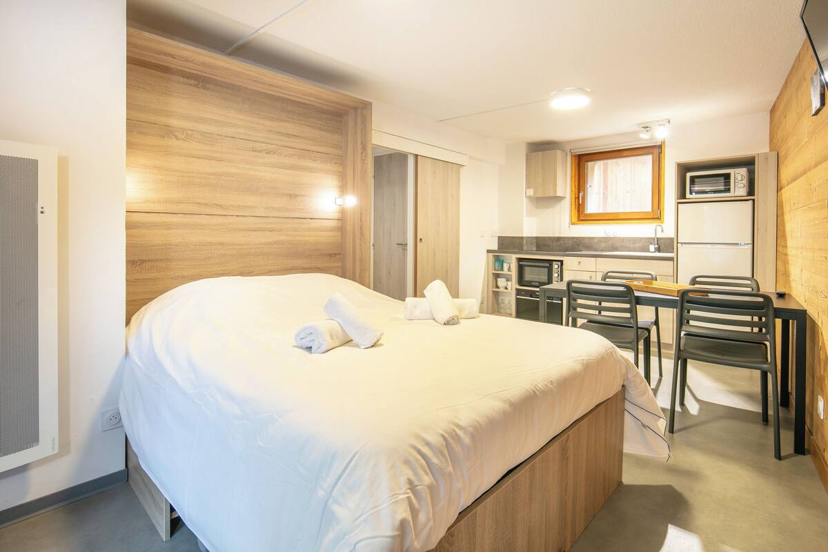 Une chambre d’hôtel avec un grand lit, une tête de lit en bois, une kitchenette équipée d’un four à micro-ondes et un coin repas avec des chaises.