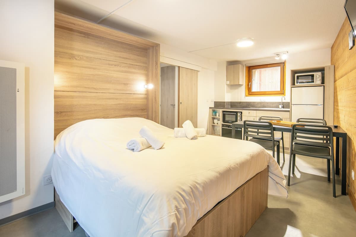 A hotel room with a large bed, a wooden headboard, a kitchenette with a microwave, and a dining area with chairs.