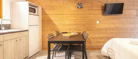 Une pièce compacte avec un mur en bois, un téléviseur à écran plat fixé au-dessus d’une table en bois avec des chaises, une petite kitchenette équipée d’un four à micro-ondes et un lit avec du linge de lit blanc.