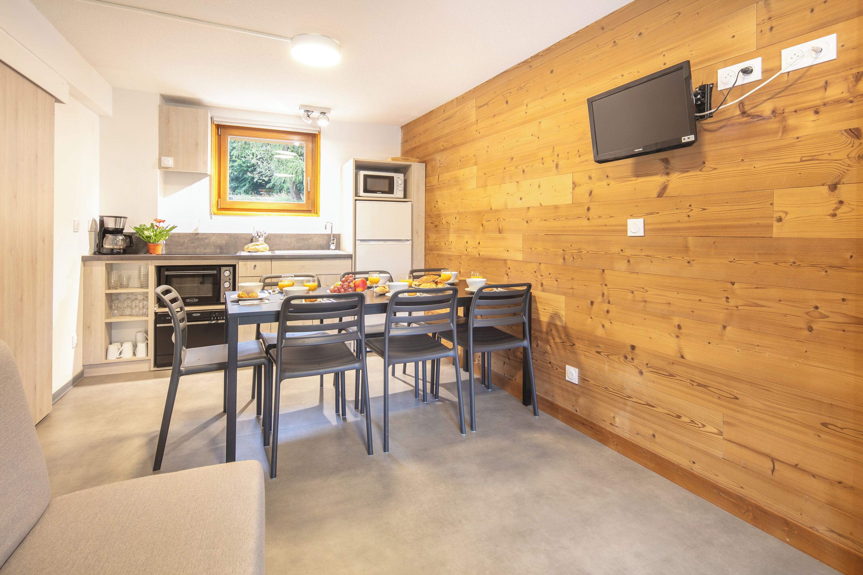 A compact kitchen with a dining area, wooden walls, and a flat-screen TV mounted above the counter.