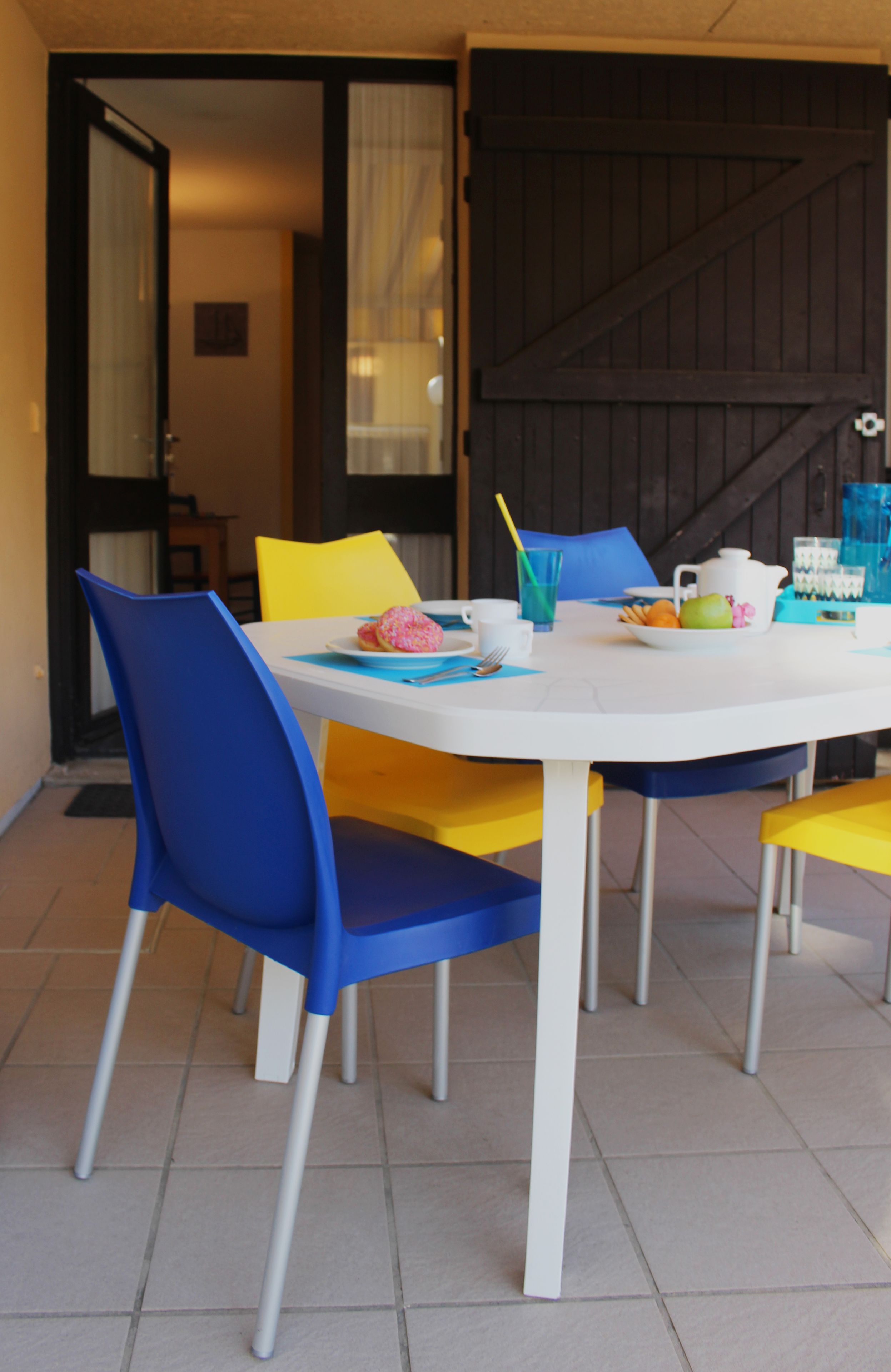 Un coin repas avec une table blanche, des chaises bleues et, au fond, un portail noir.