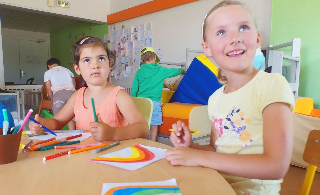 Two children drawing at a table with art supplies, a whiteboard with children's drawings in the background, and a green wall.