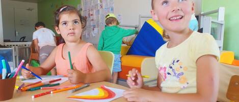 Two children drawing at a table with art supplies, a whiteboard with children's drawings in the background, and a green wall.