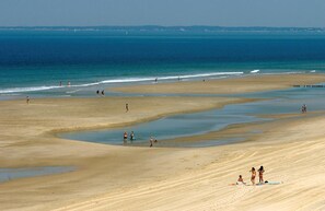 A wide sandy beach with calm water, a few people walking, and some sitting on the sand.