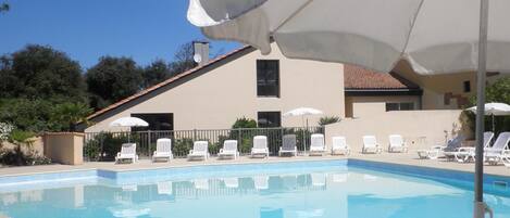 Une piscine entourée de chaises longues blanches et de parasols, le tout bordé d’un bâtiment doté d’un toit en tuiles et d’une clôture.