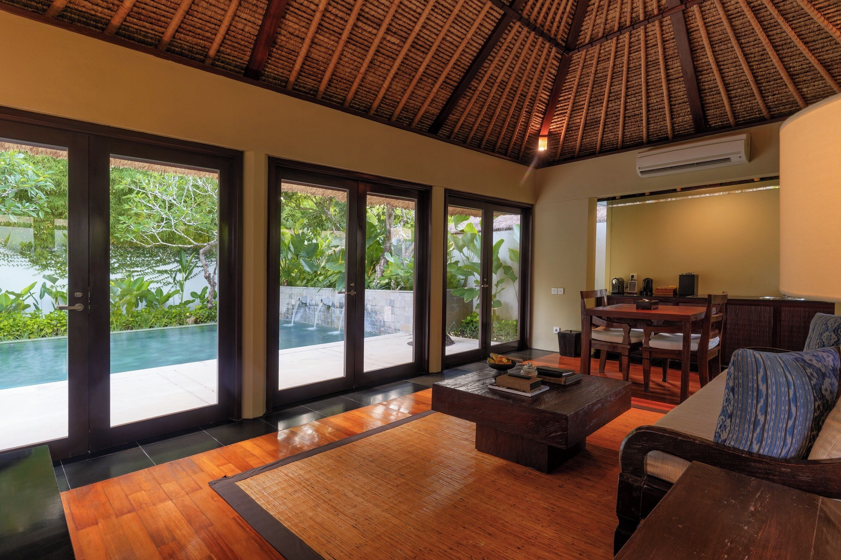 A spacious living area with a large wooden coffee table, a sofa, and a dining table with chairs. Large sliding glass doors lead to a pool area with lush greenery.