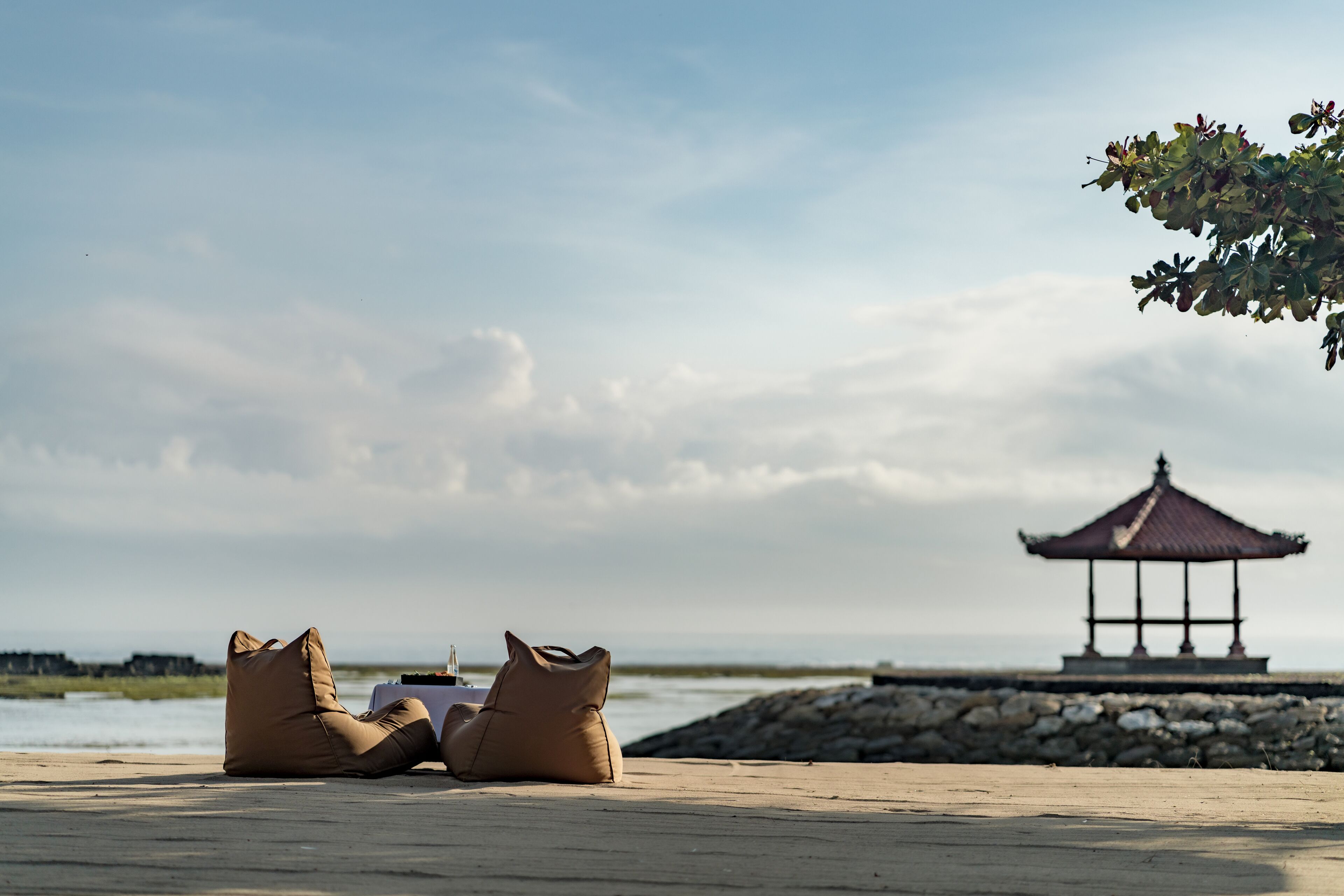 A beach setting with two bean bags and a table with a bottle, a gazebo in the background, and a stone wall.