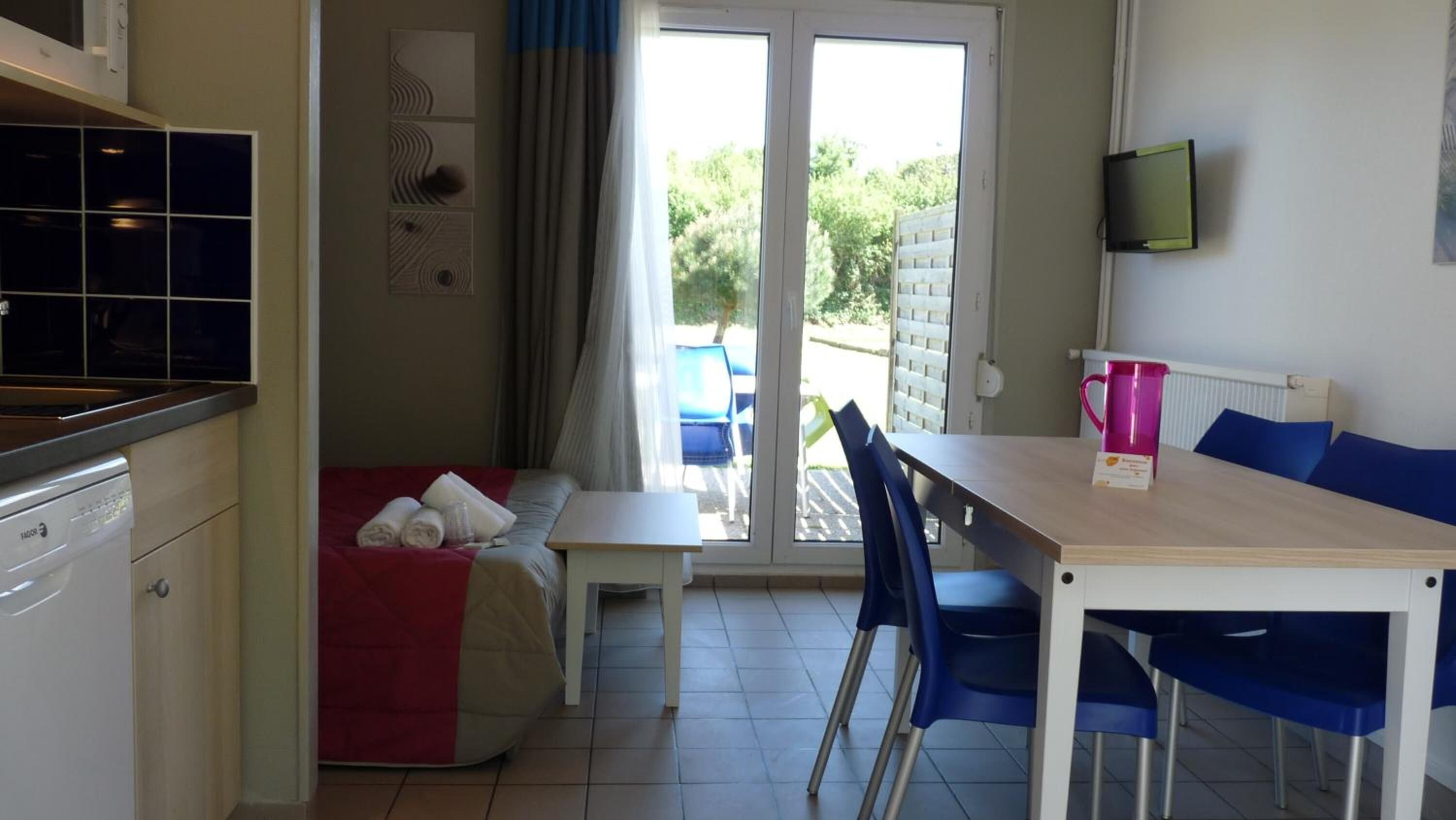 A compact room with a kitchenette, a bed with a red blanket, a table with chairs, and a TV on the wall.