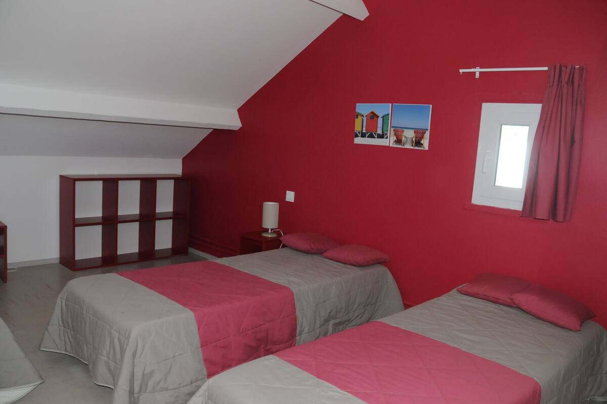 A room with two beds, a red wall, a window with curtains, and a small table with a lamp.