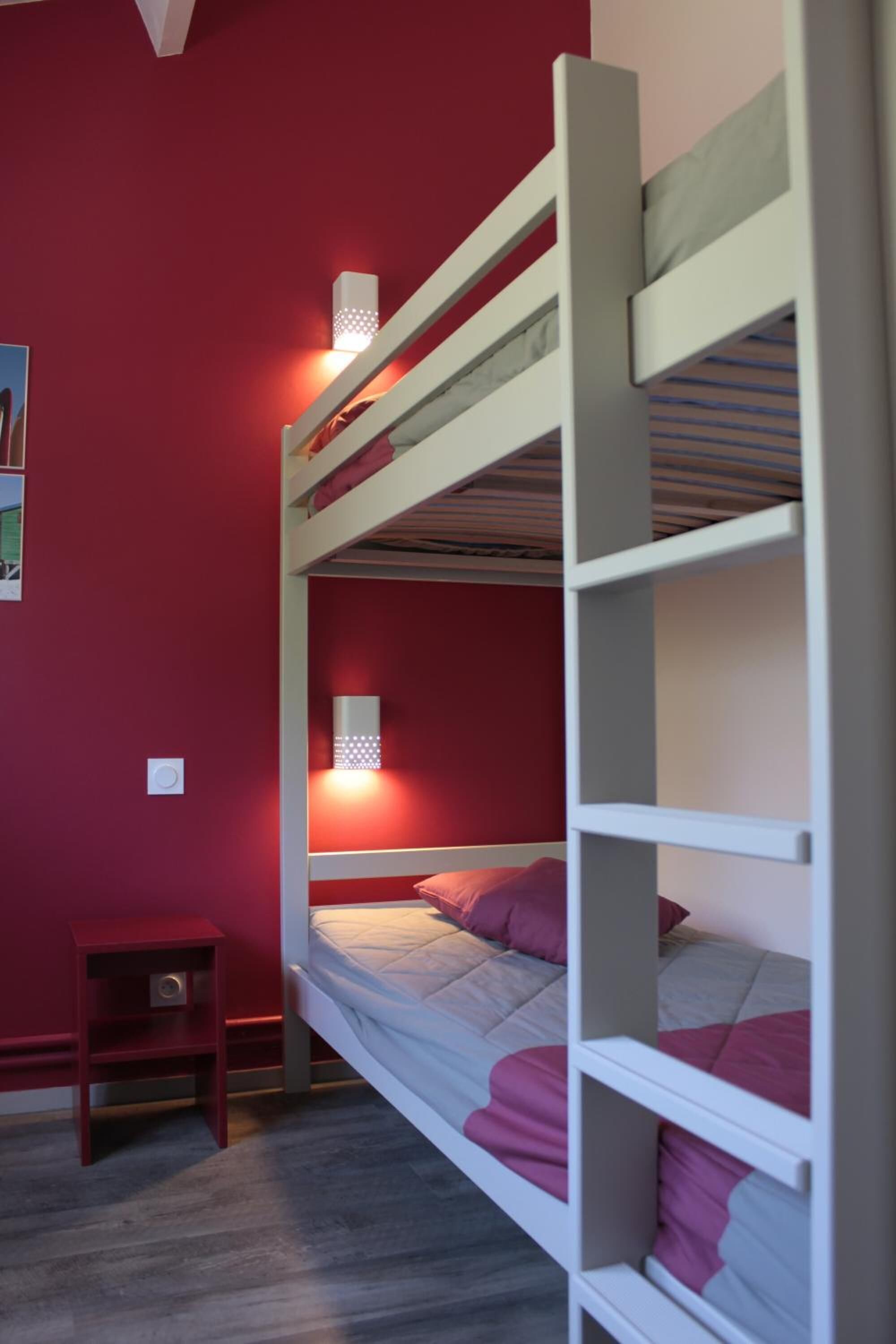 Een slaapkamer met een stapelbed, een rode wand, een klein houten nachtkastje en twee wandlampen.