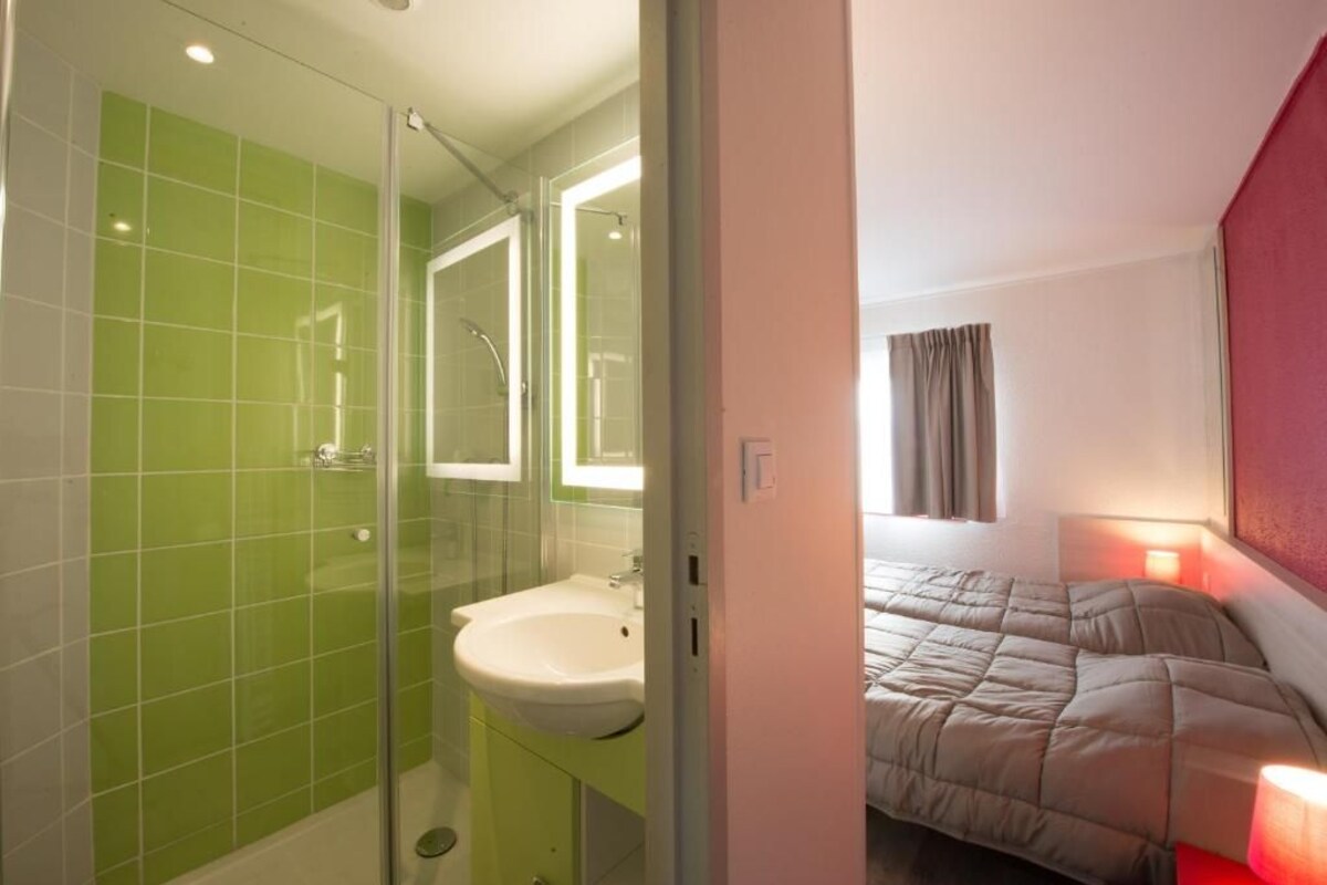 A bathroom with green tiles, a glass shower enclosure, a white sink, and a bed with a red lamp.