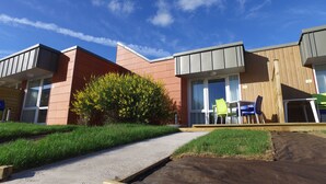 A modern building with a mix of brick and wood paneling, large windows, and a small garden area with a pathway leading to outdoor seating. - VVF Blériot-Plage (Sangatte)