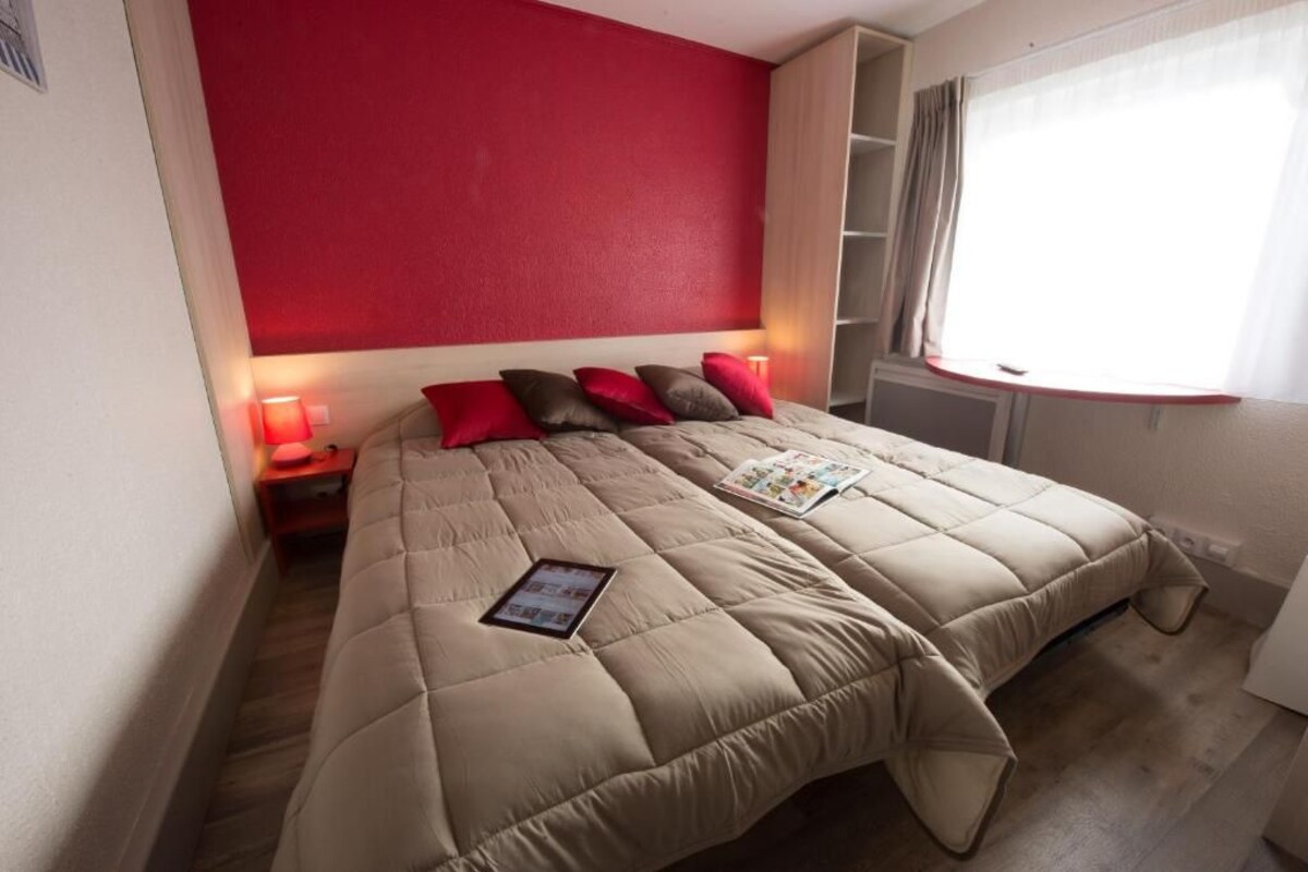 A neatly made bed with a red wall behind it, a bedside table with a lamp, and a window with curtains.