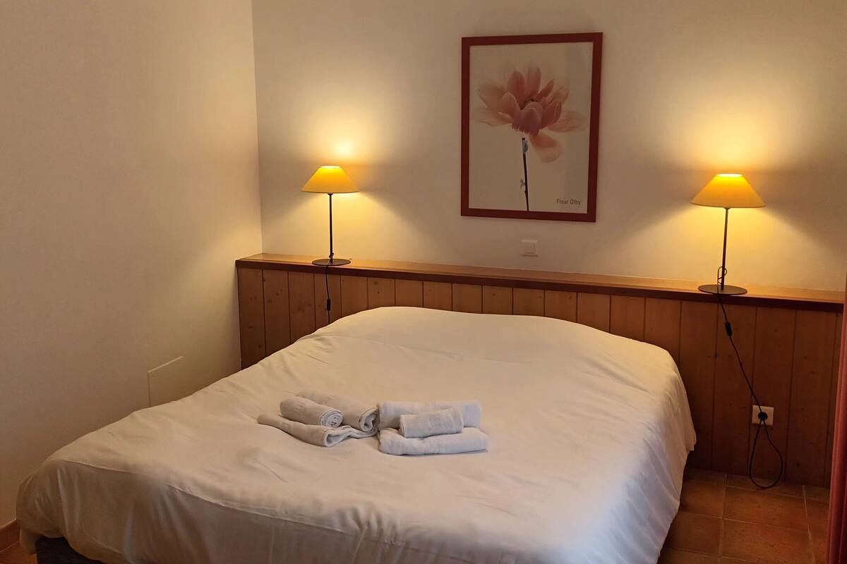 Ein ordentlich bezogenes Bett mit Handtüchern, zwei Nachttischlampen, einem gerahmten Bild an der Wand und einer Deckenleuchte.