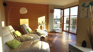 A modern living room with a wooden wall, a sofa with green cushions, a side table with a vase, and a large window offering a view of the outdoors.