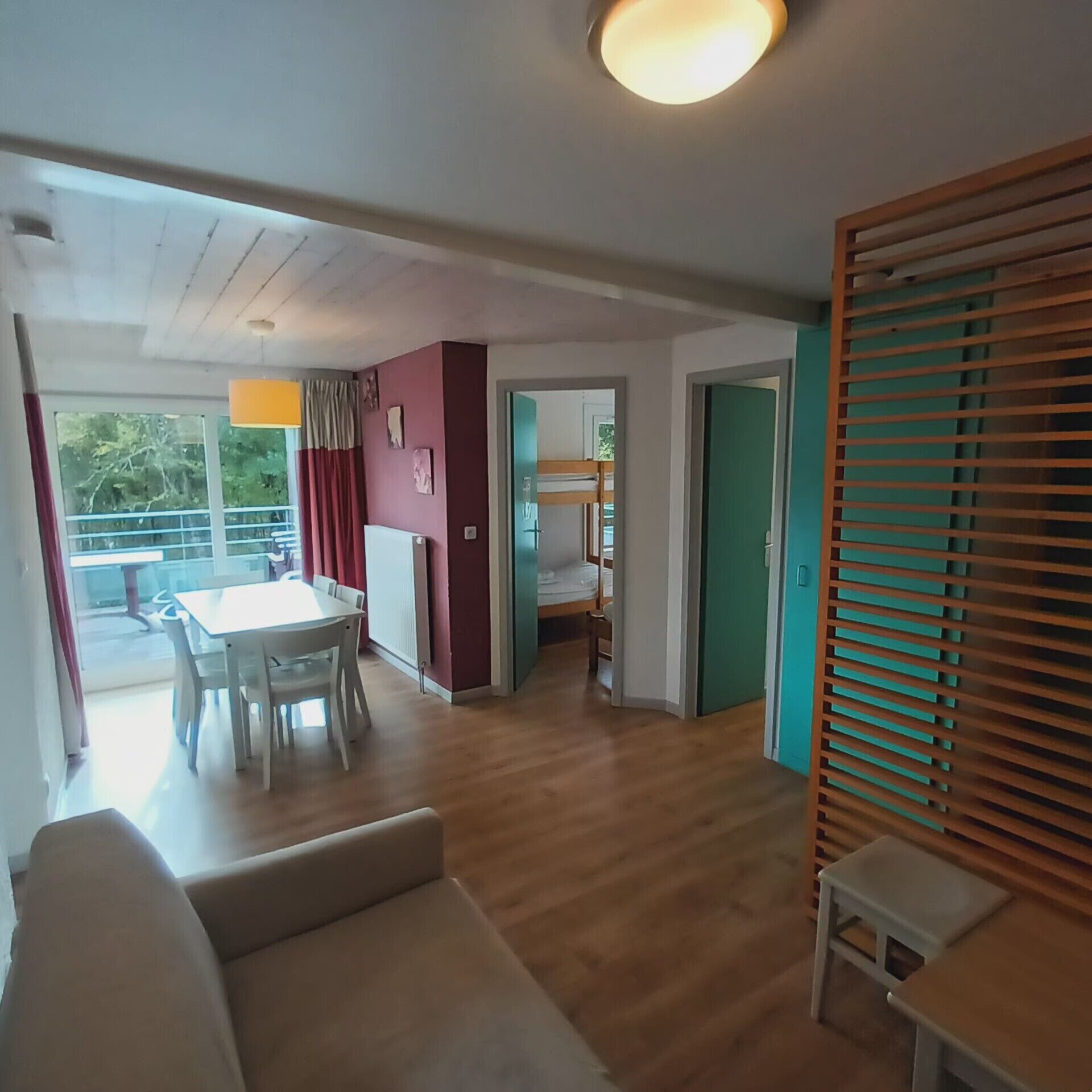A room with a wooden floor, a white sofa, a dining table with chairs, a bunk bed, and a sliding door with blinds.