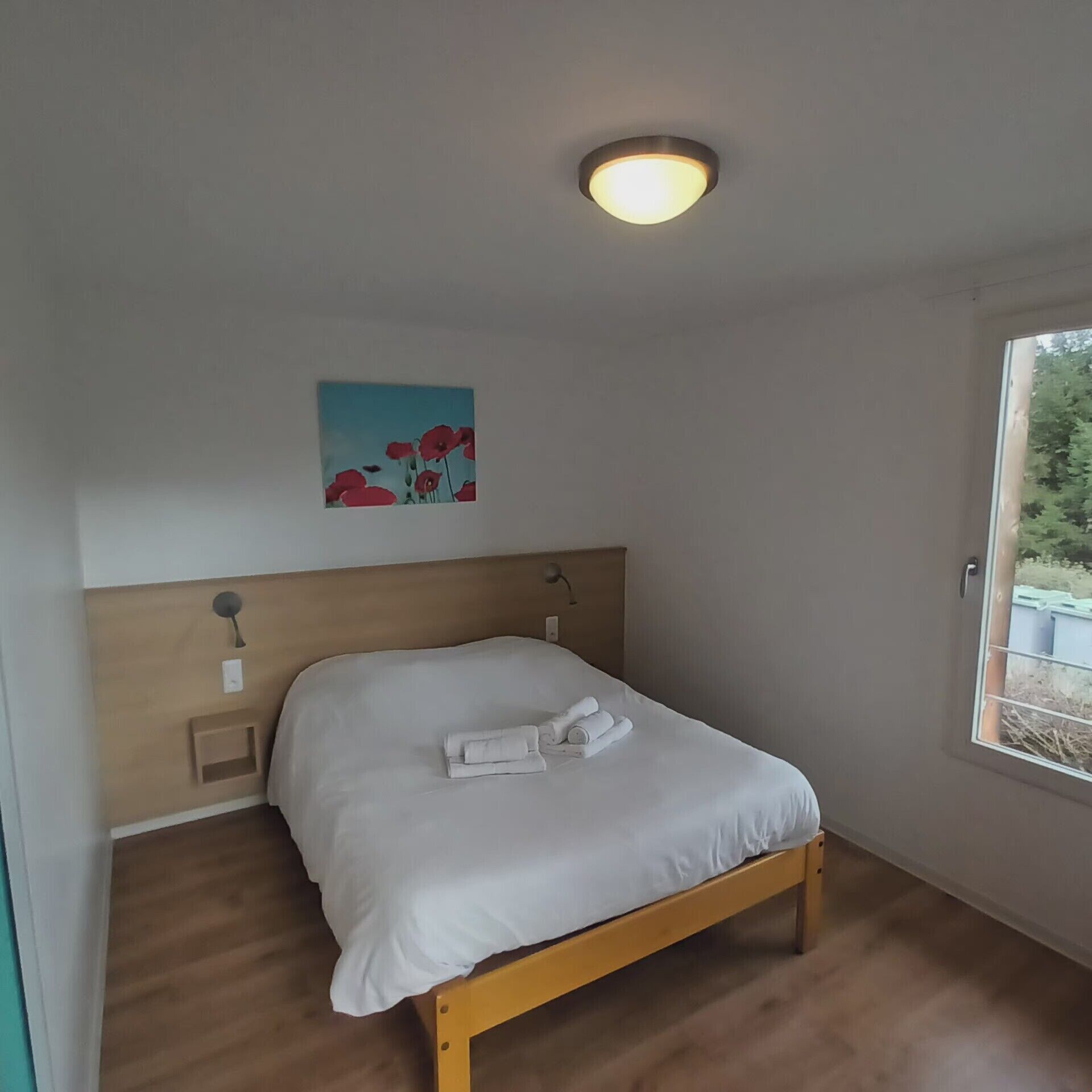 Een enkel bed met houten frame, een wit matras en netjes opgevouwen handdoeken. Er is een plafondlamp en een wandlamp. Op de muur hangt een schilderij van rode bloemen.