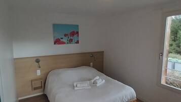 Een enkel bed met houten frame, een wit matras en netjes opgevouwen handdoeken. Er is een plafondlamp en een wandlamp. Op de muur hangt een schilderij van rode bloemen.