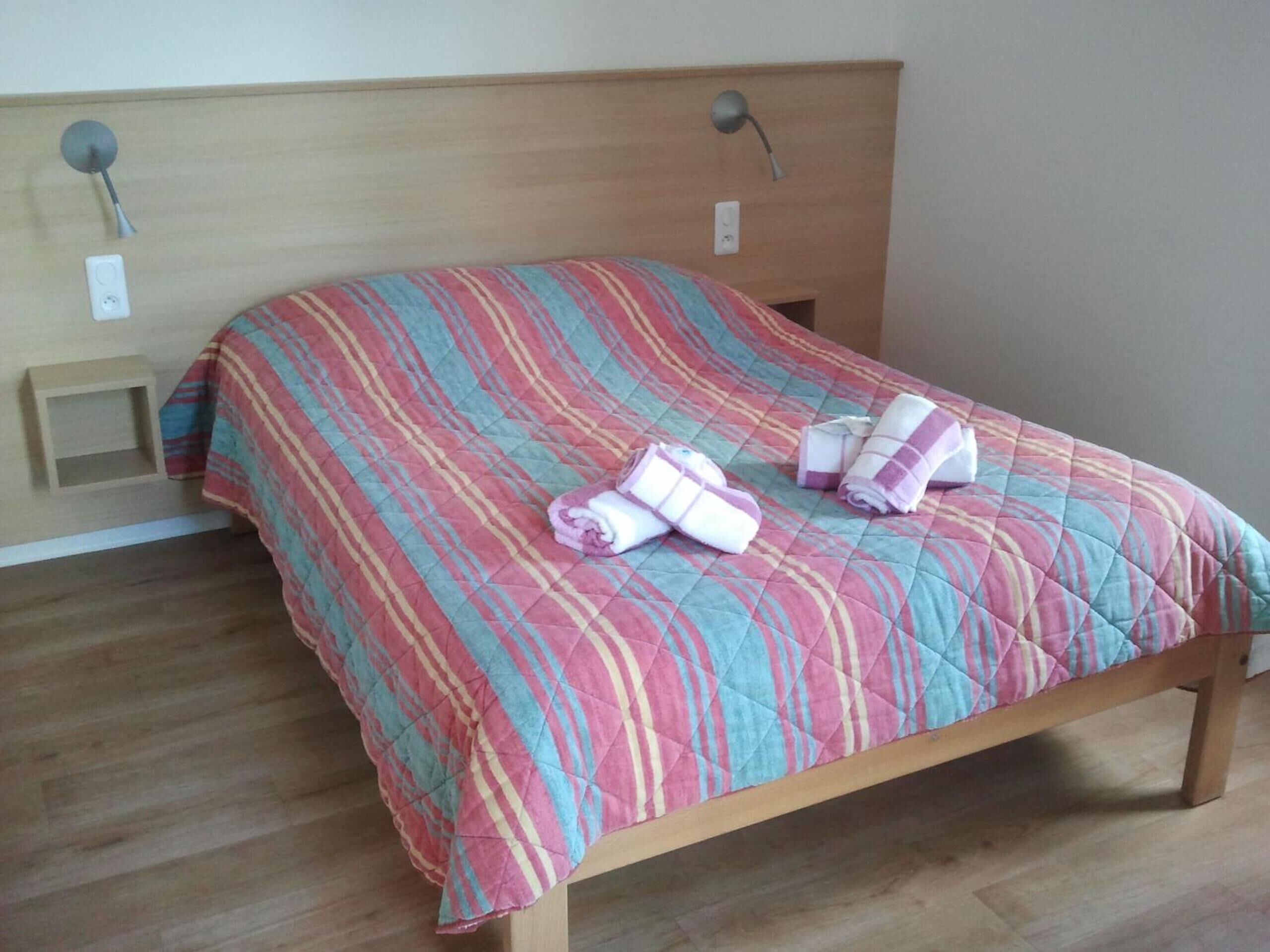 A bed with a striped quilt and rolled towels, a wooden headboard, and a bedside table with a lamp.