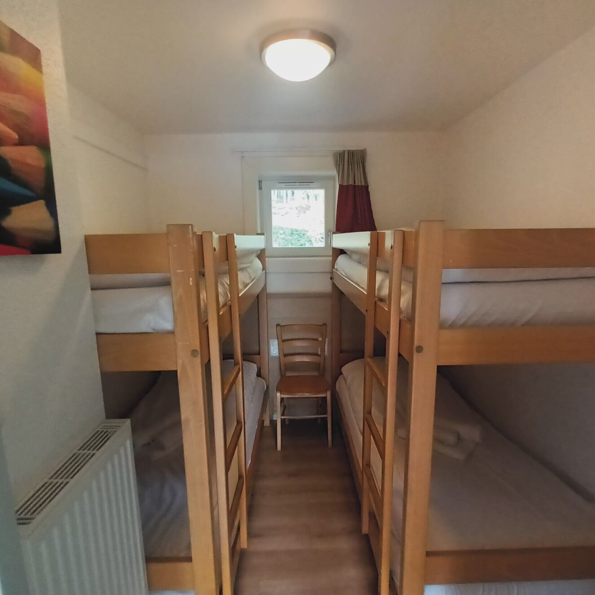 Un dormitorio con literas, tres literas de madera, una ventana pequeña con cortinas y un radiador.