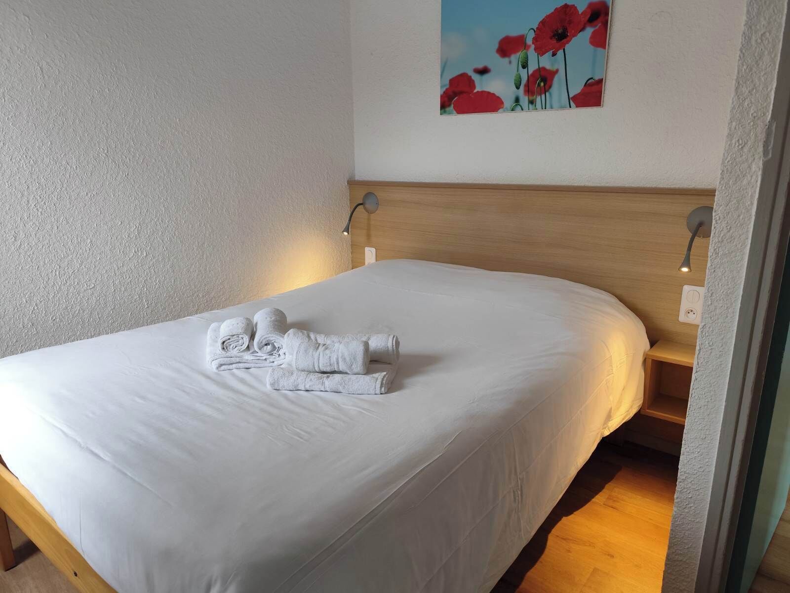 A neatly made bed with white linens and rolled towels, a wooden headboard, and a painting of red flowers on the wall.